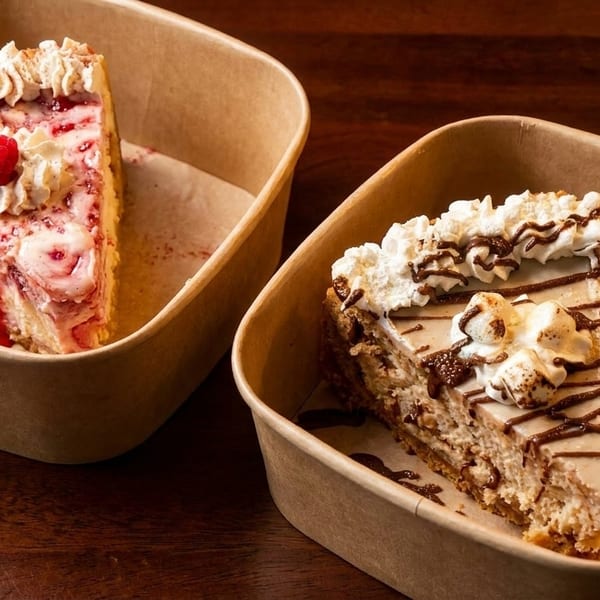 Two slices of cheesecake in takeout containers, one raspberry and one chocolate with whipped cream.