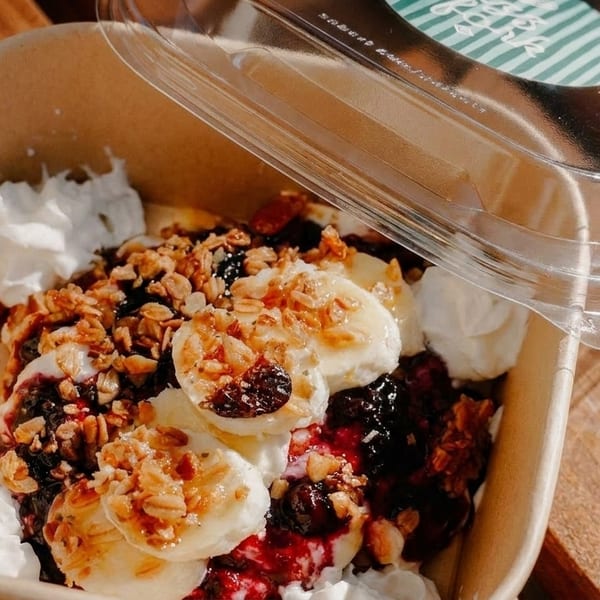 A takeout container filled with yogurt, granola, banana slices, berries, and whipped cream.
