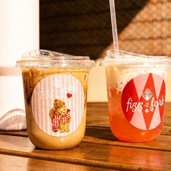 Two Fizz & Fork drinks, one iced coffee and one red beverage, on a wooden table outdoors.