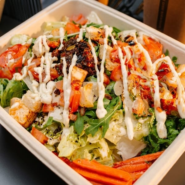A vibrant salad with chicken, tomatoes, and dressing in a compostable container on a dark table.