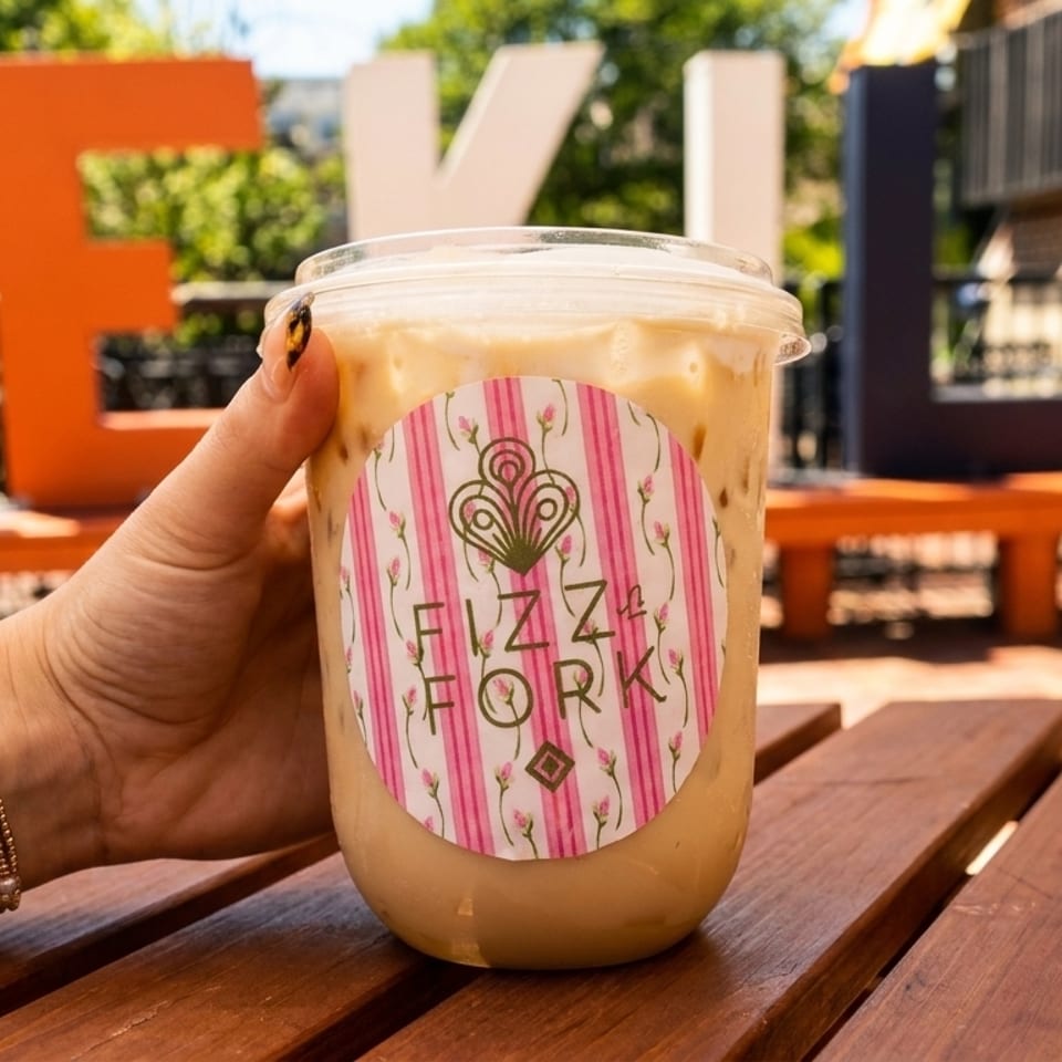 Hand holding an iced coffee with a "Fizz & Fork" logo, on a wooden table with large letters in the background.