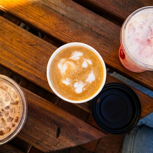 Overhead view of three drinks on a wooden table: an iced coffee, a latte with art, and a pink iced drink.